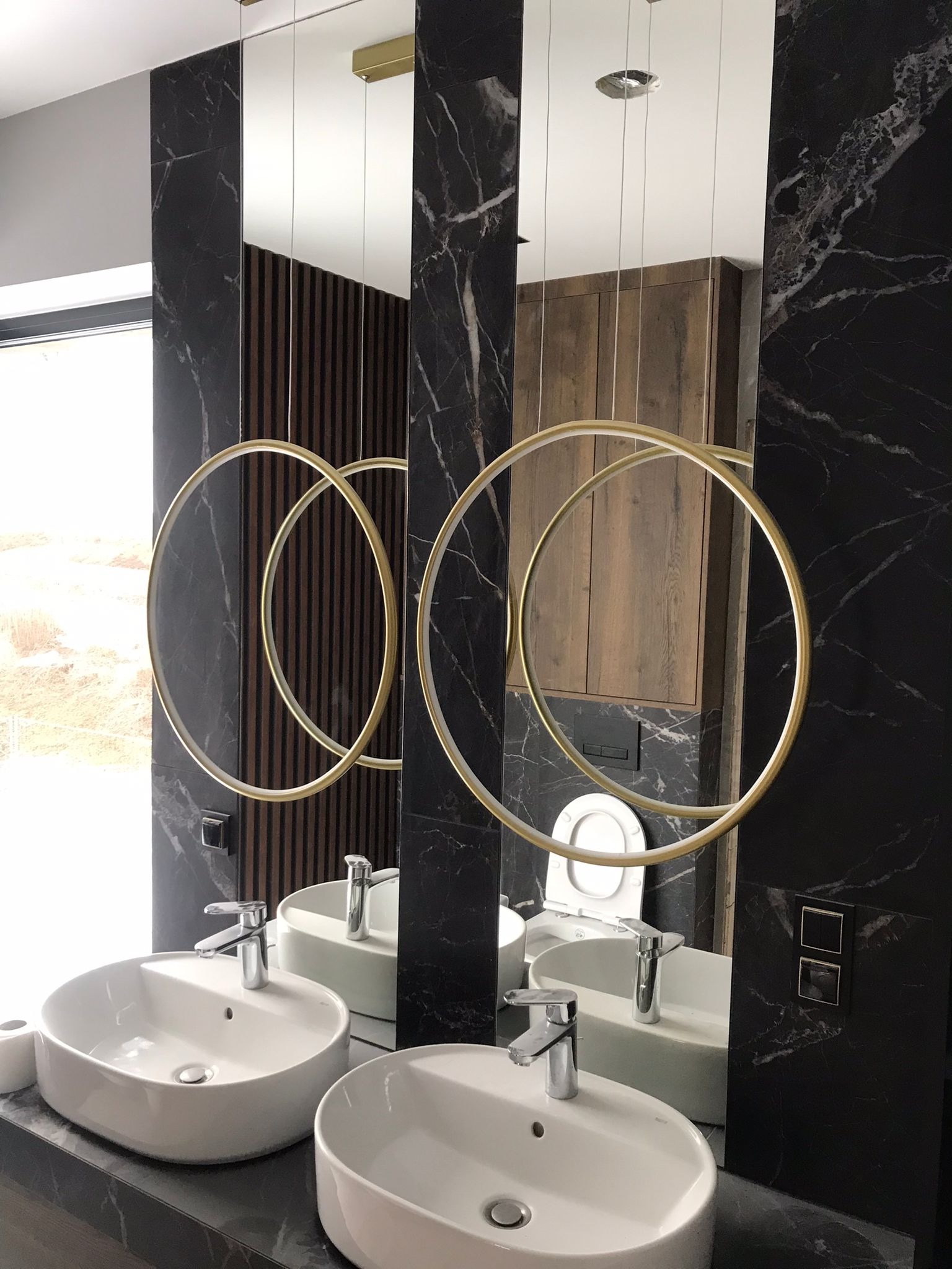 Nowoczesna łazienka z dwoma okrągłymi lustrami, białymi umywalkami nablatowymi, czarnymi marmurowymi ścianami i drewnianymi akcentami.