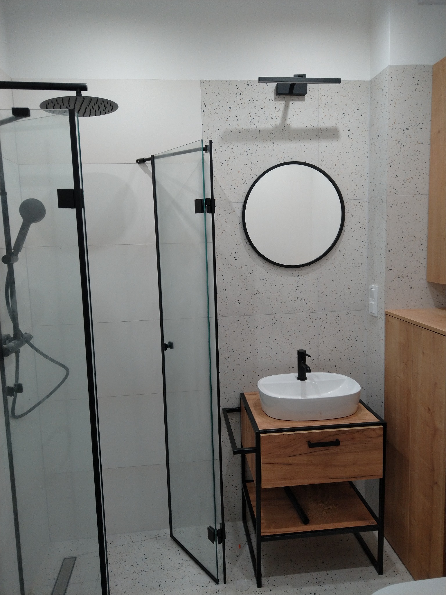Nowoczesna łazienka z kabiną prysznicową typu walk-in z czarnymi ramami, okrągłym lustrem nad umywalką nablatową na szafce z drewnianym blatem i czarnymi metalowymi nogami, oraz płytkami...