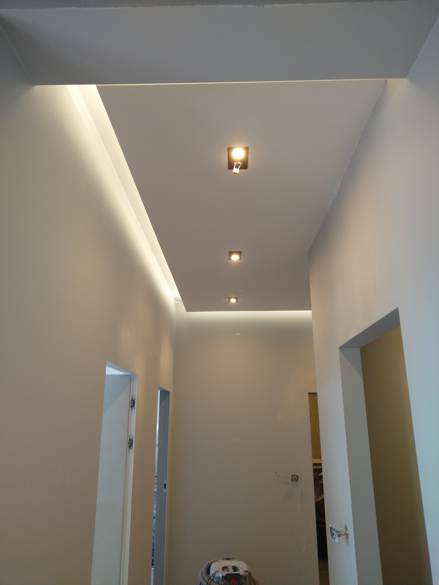 Nowoczesny korytarz z podwieszanym sufitem z oświetleniem LED i trzema punktowymi lampami, widok w perspektywie, z widocznymi elementami instalacji elektrycznej i sprzętem budowlanym w tle.