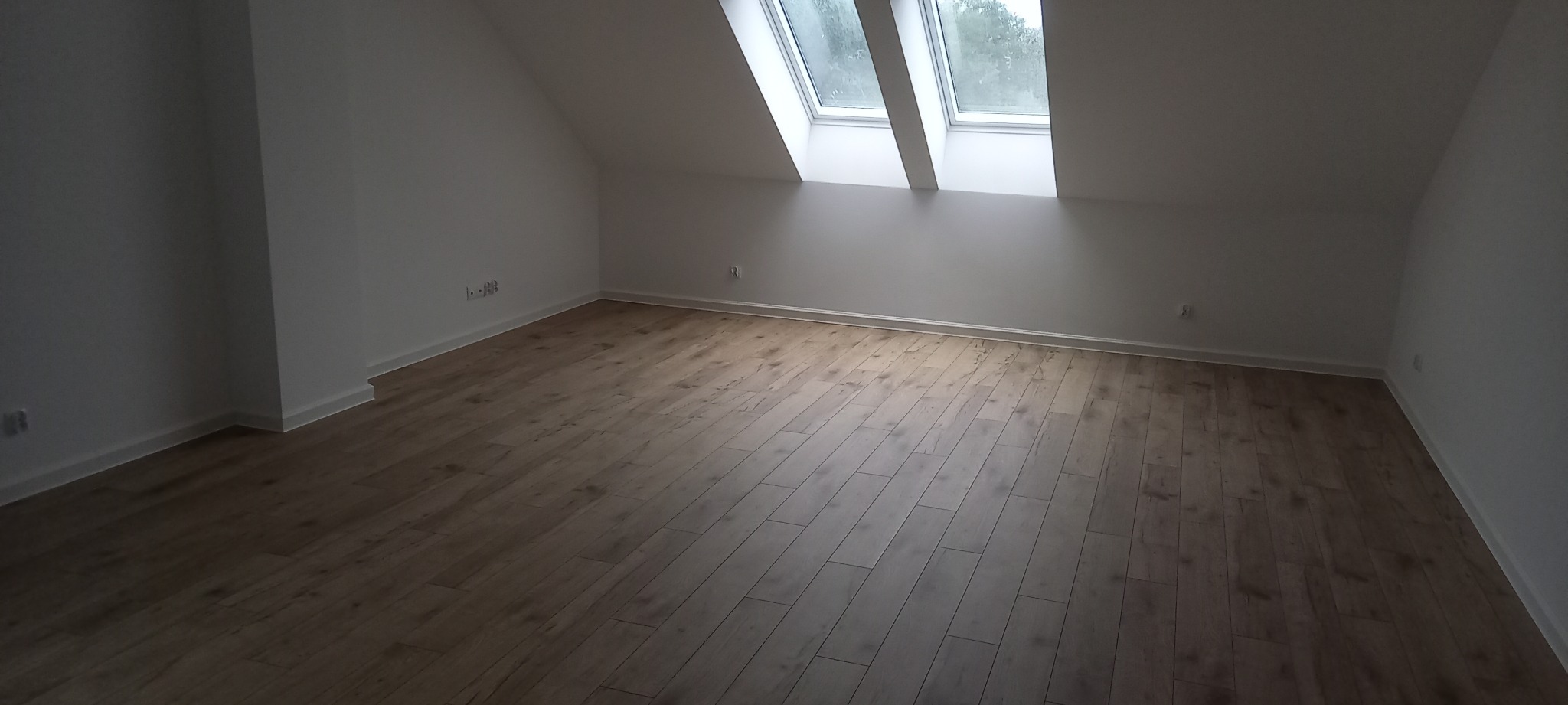Poddasze z nową, jasną podłogą laminowaną, widoczne dwa okna dachowe i białe ściany.