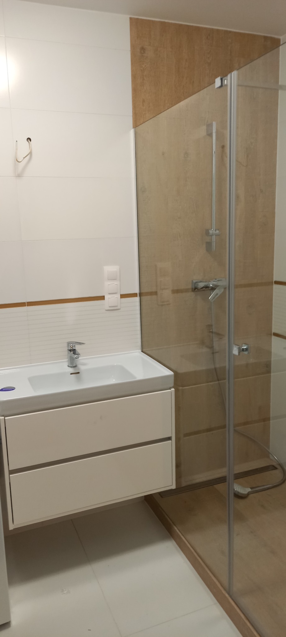 Nowoczesna łazienka z białą umywalką z szafką, kabiną prysznicową ze szklaną ścianką i panelami imitującymi drewno na ścianie, widoczne białe płytki i chromowana armatura.