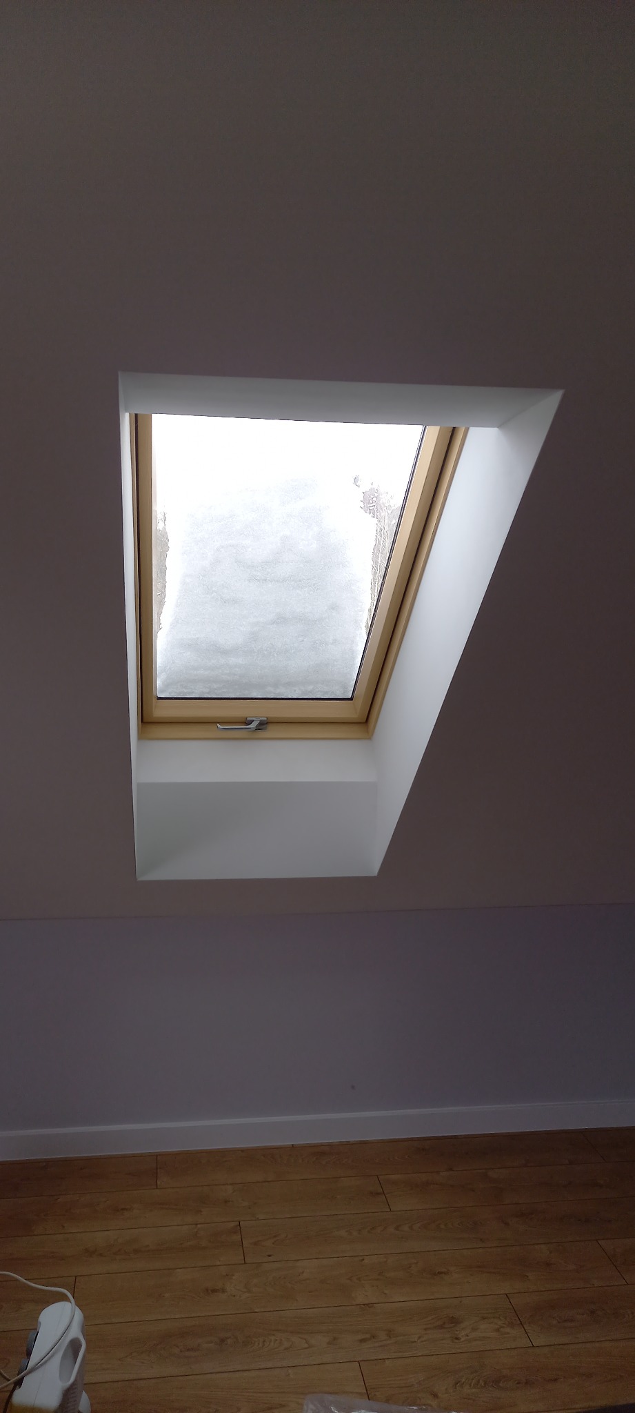 Okno dachowe z widokiem na zaśnieżony krajobraz, osadzone w białej wnęce na poddaszu, z widocznymi panelami podłogowymi.