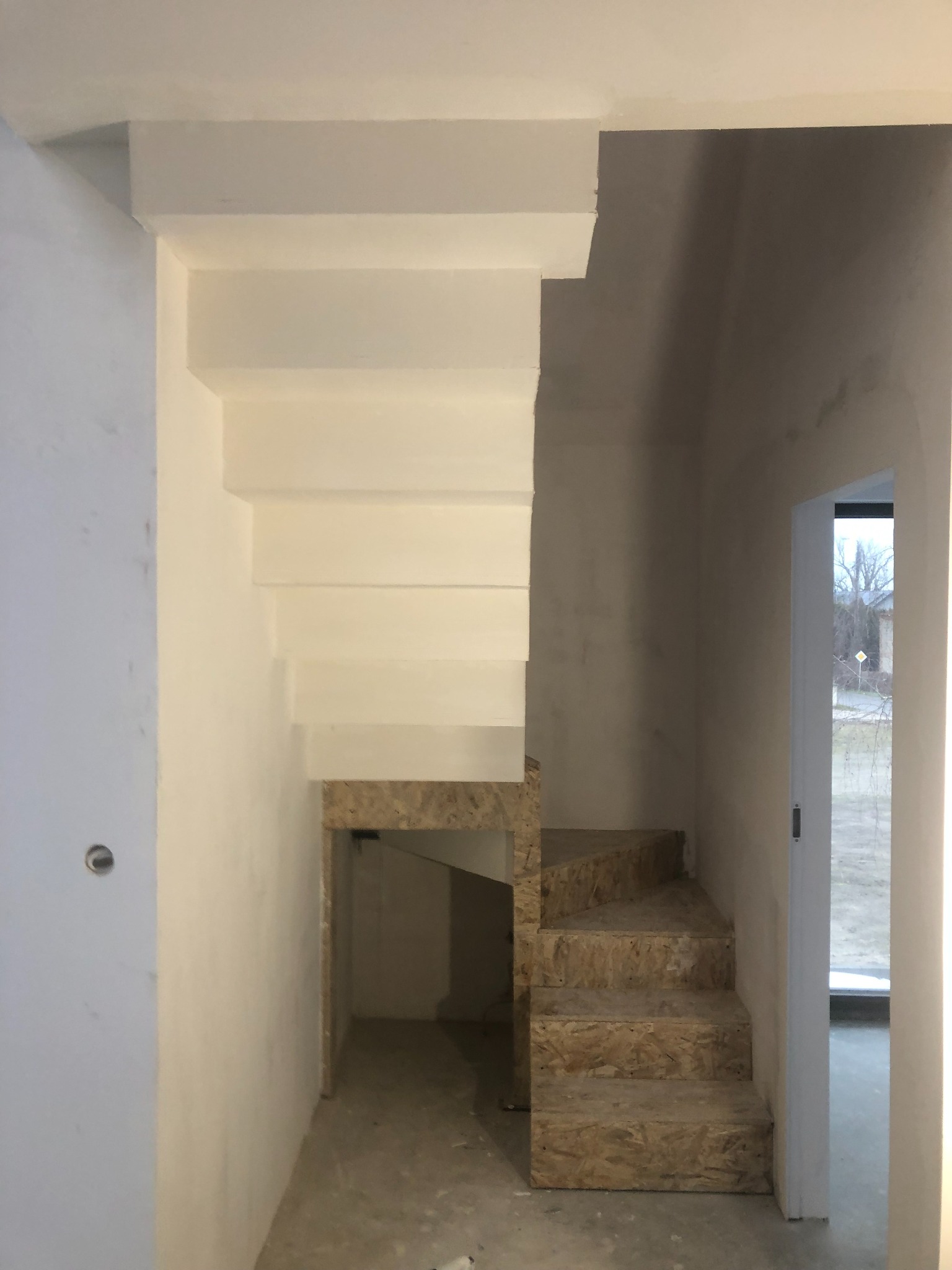 Surowe schody z płyty OSB w trakcie budowy, z widoczną konstrukcją pod schodami i fragmentem ściany z gładzią, widok na zewnątrz przez otwarte drzwi.