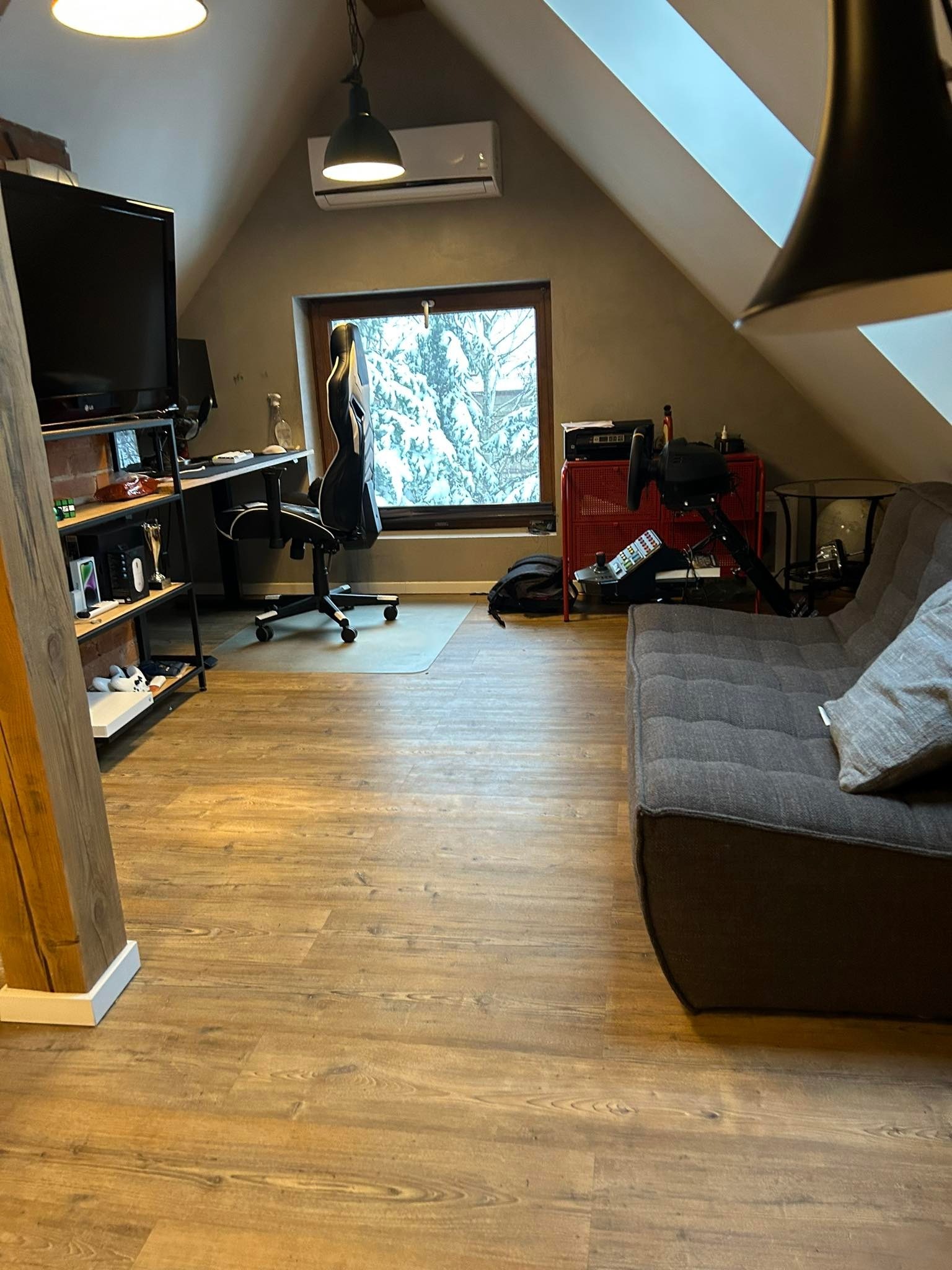 Pokój na poddaszu z drewnianą podłogą, biurkiem, fotelem gamingowym i sofą, widok na zaśnieżone drzewa za oknem