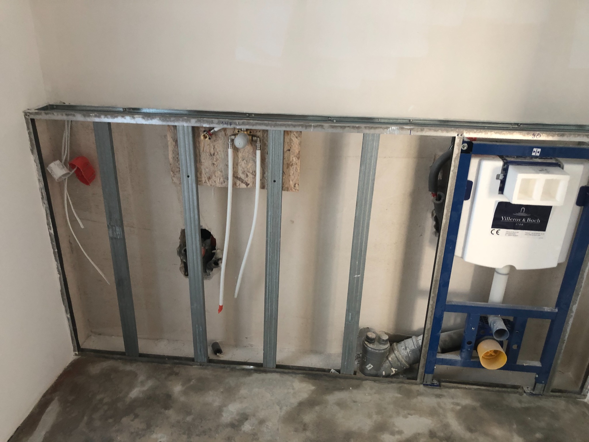 Stelaż podtynkowy Villeroy & Boch zamontowany w konstrukcji z profili stalowych, widoczne instalacje wodno-kanalizacyjne w surowym stanie, przygotowanie do obudowy ściany.