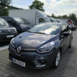 Szary Renault Clio kombi zaparkowany na brukowanym placu, widoczny z przodu, z innymi samochodami i białym busem w tle, tablica rejestracyjna WW 906SF.