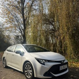 Click&Car - Biała Toyota Corolla kombi na leśnej drodze, jesienne drzewa w tle. Auto z widoczną tablicą rejestracyjną, zaparkowane w naturalnym otoczeniu.