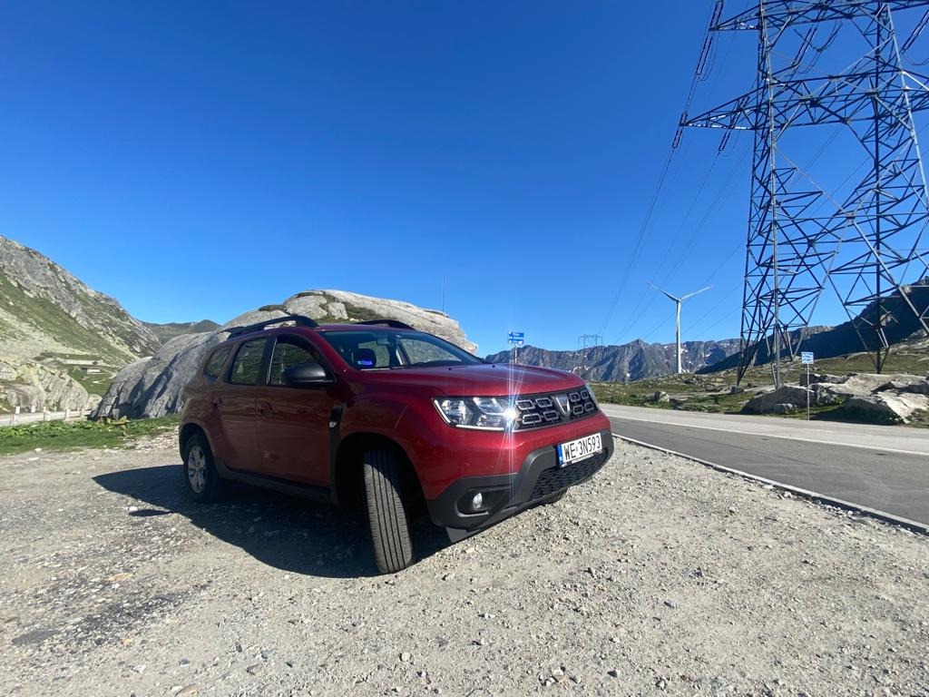 Czerwona Dacia Duster 2022 zaparkowana na żwirowym poboczu z widokiem na góry, turbiny wiatrowe i linię wysokiego napięcia w tle.