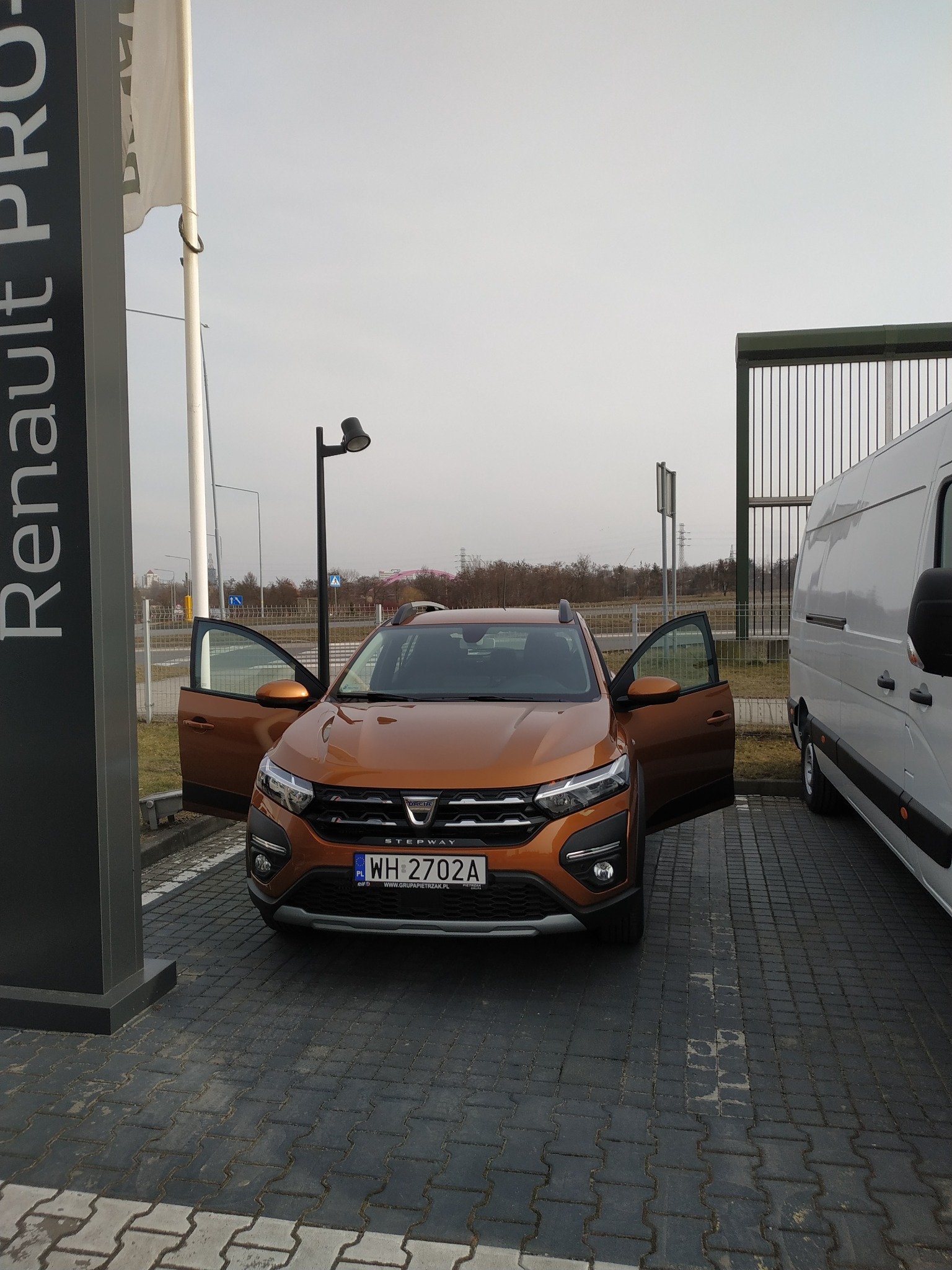 Pomarańczowa Dacia Sandero Stepway 2022 z otwartymi drzwiami, zaparkowana na kostce brukowej obok znaku Renault Pro i białego busa.