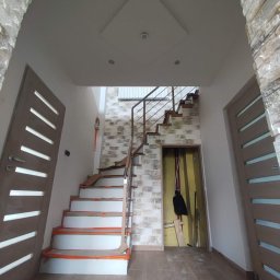 Alan Sobieraj - Wnętrze w trakcie prac wykończeniowych: zabezpieczone schody z metalową barierką, dwie nowoczesne, drewniane drzwi z przeszkleniami po bokach, dekoracyjna ściana z imitacji kamienia, sufit...