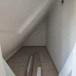 Poddasze ze skosami przed pracami wykończeniowymi, widoczne płyty gipsowo-kartonowe oparte o ścianę, surowa żarówka zwisająca z sufitu.