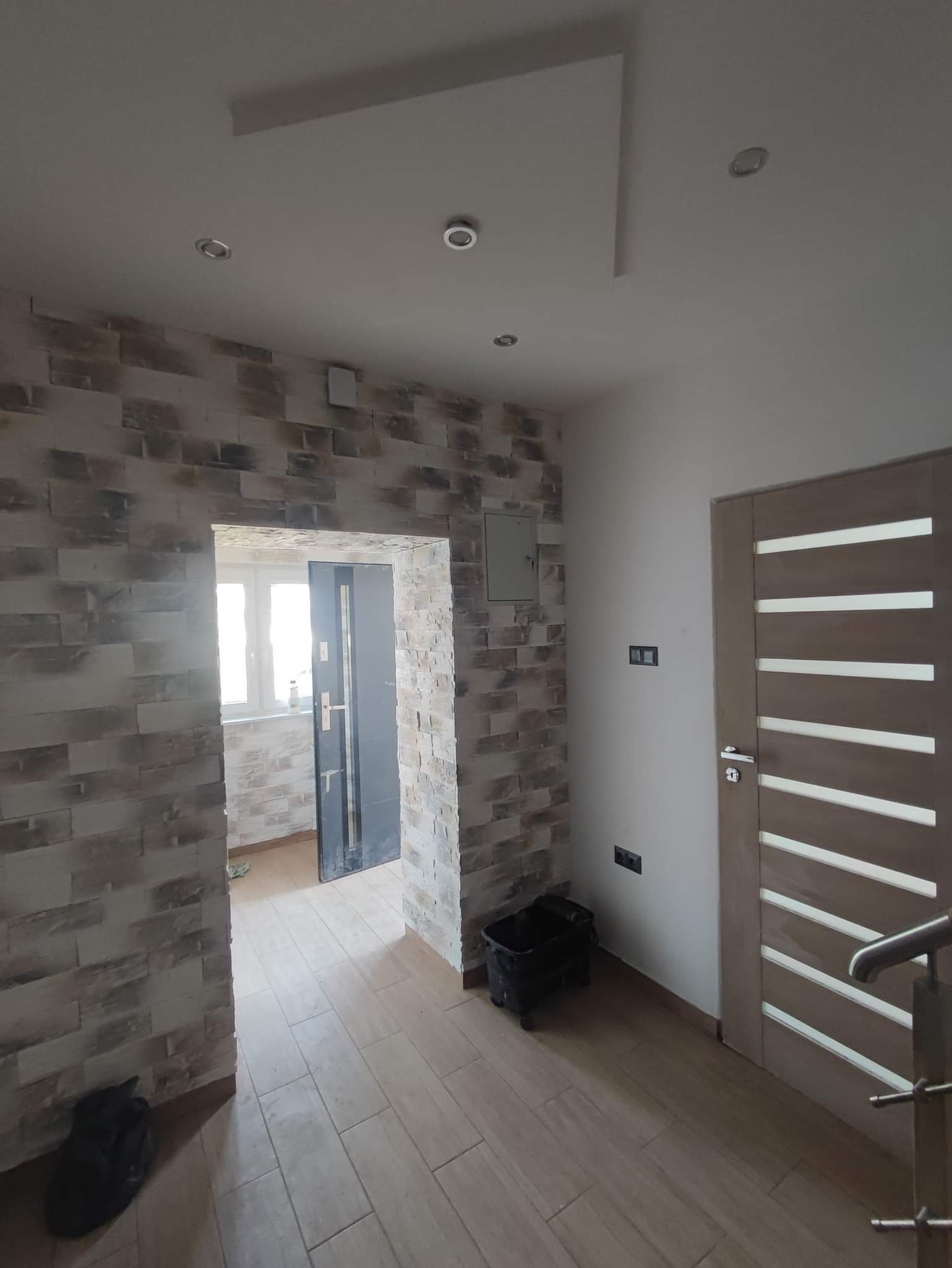 Wykończone wnętrze mieszkania z dekoracyjną ścianą z kamienia imitującego cegłę, drewnianą podłogą, świeżo zamontowanymi drzwiami wewnętrznymi z poziomymi przeszkleniami i czarnym wiadrem...