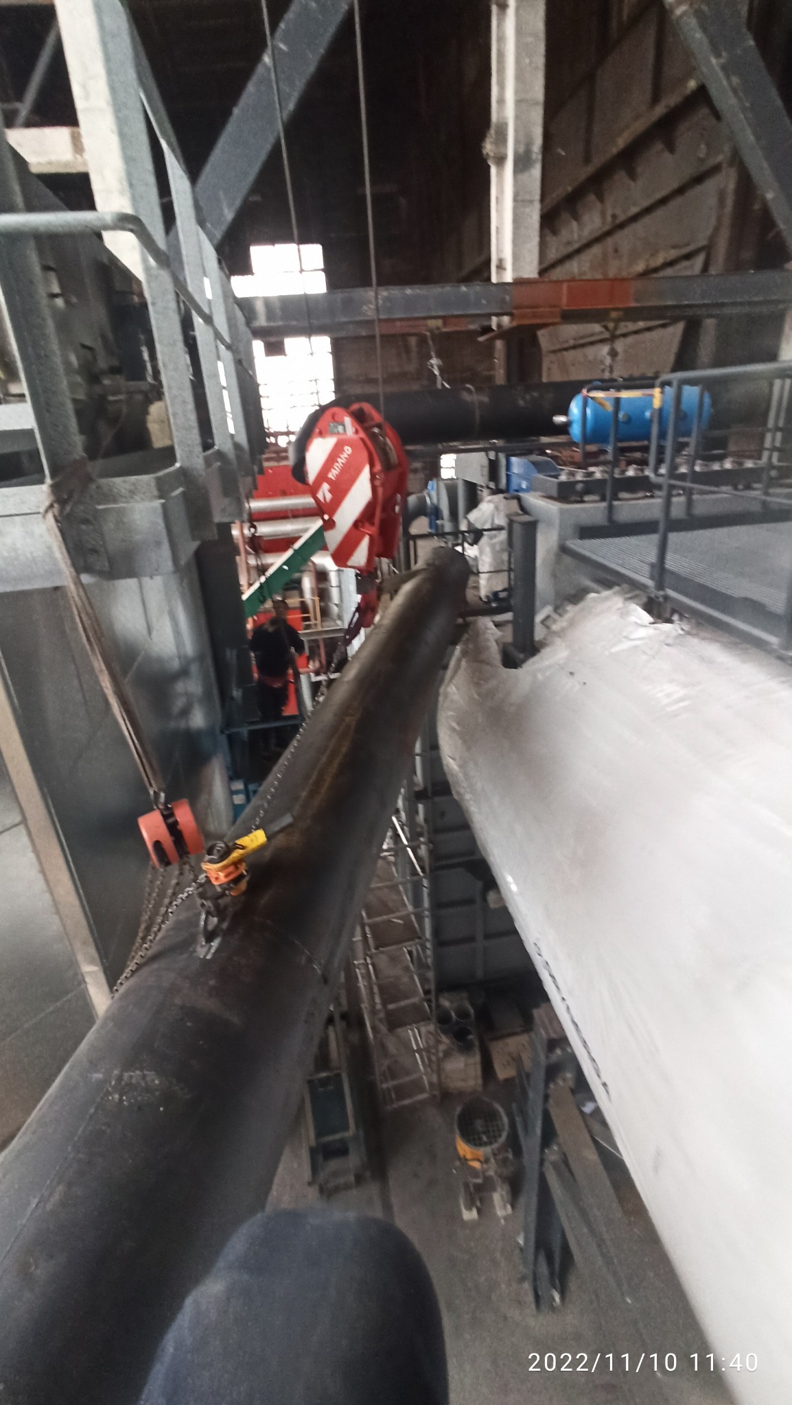 Widok z góry na fragment konstrukcji hali z rurami, w trakcie montażu, z użyciem czerwonego wciągnika łańcuchowego marki Tadano, z pracownikiem w tle.