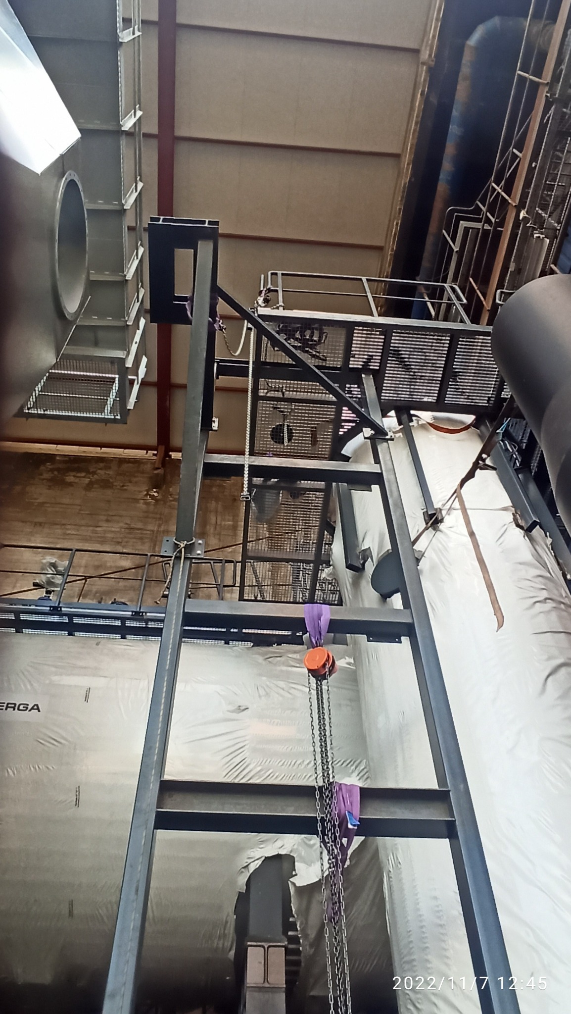 Wnętrze hali w trakcie budowy, widoczne stalowe konstrukcje, elementy wentylacyjne i podwieszony łańcuch z pomarańczowym bloczkiem.