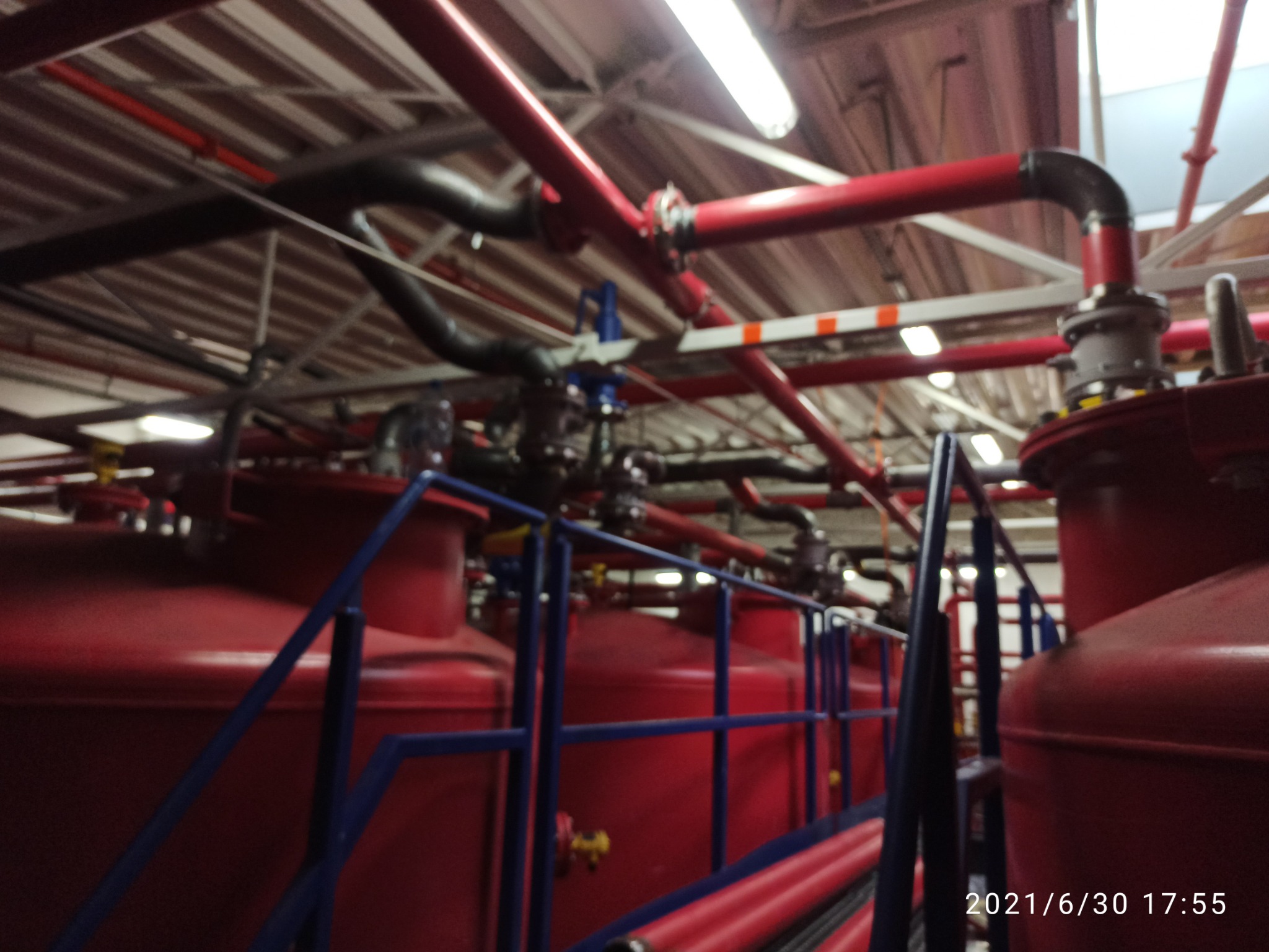 Wnętrze hali przemysłowej z widocznymi czerwonymi zbiornikami, rurami i niebieskimi barierkami ochronnymi, oświetlone lampami jarzeniowymi