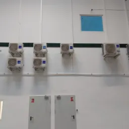 Rząd ośmiu klimatyzator&oacute;w Haier zamontowanych na białej ścianie w hali produkcyjnej, widoczne drzwi przeciwpożarowe i okno z niebieską folią.