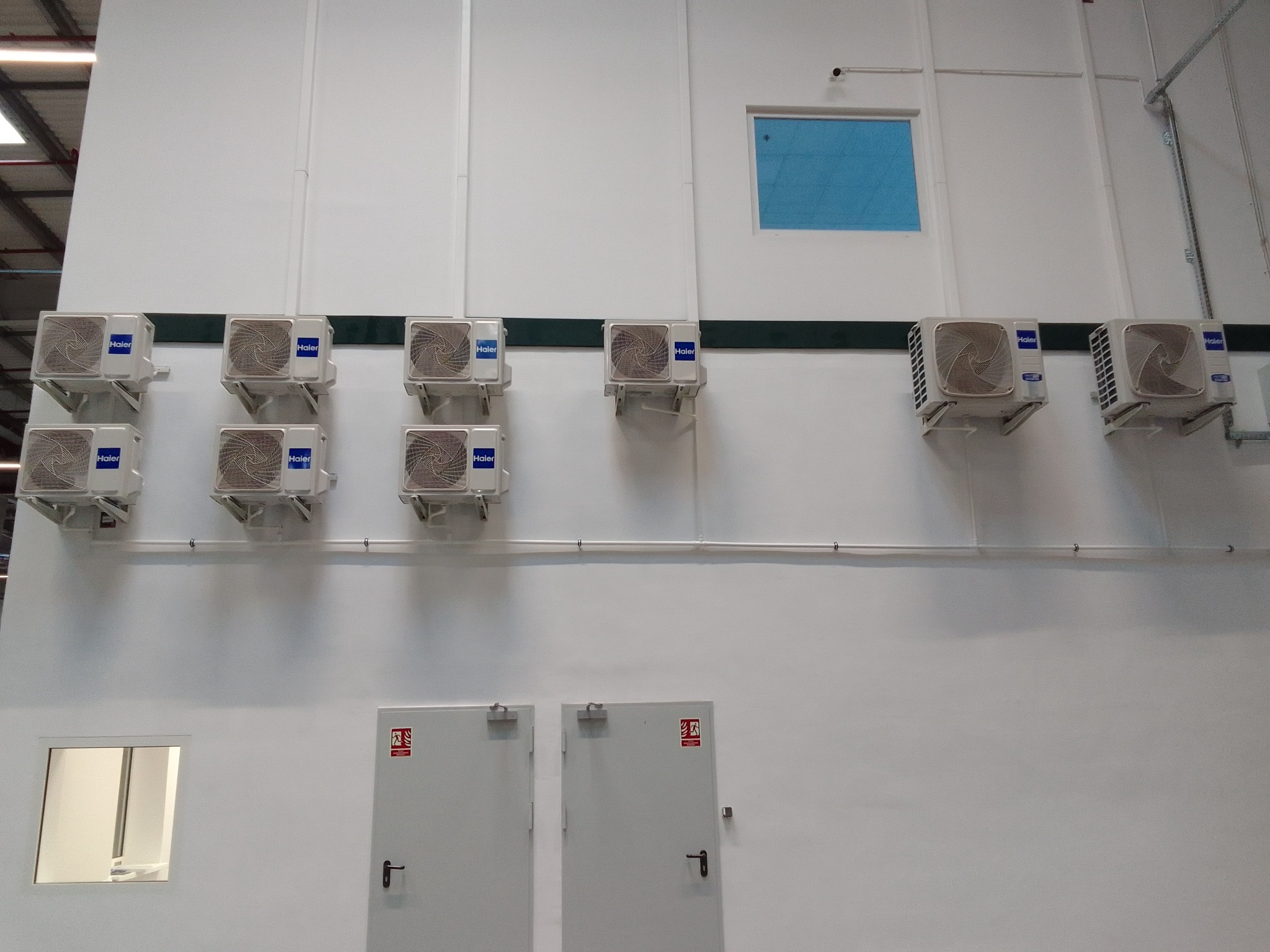 Rząd ośmiu klimatyzatorów Haier zamontowanych na białej ścianie w hali produkcyjnej, widoczne drzwi przeciwpożarowe i okno z niebieską folią.