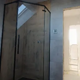 Kabina prysznicowa z czarnymi ramami w łazience z szarymi i drewnopodobnymi płytkami, widoczny fragment okna dachowego i drabina w przejściu do innego pomieszczenia.