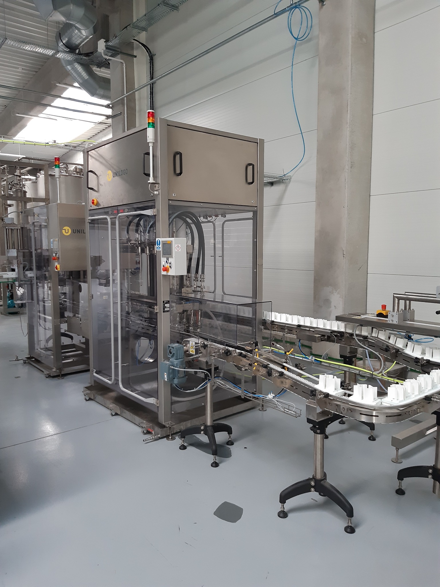 Automatyczna linia produkcyjna z transporterem i modułami do pakowania, widoczne białe pojemniki na taśmie, szare ściany i podłoga hali, logo Unilogo na maszynie.