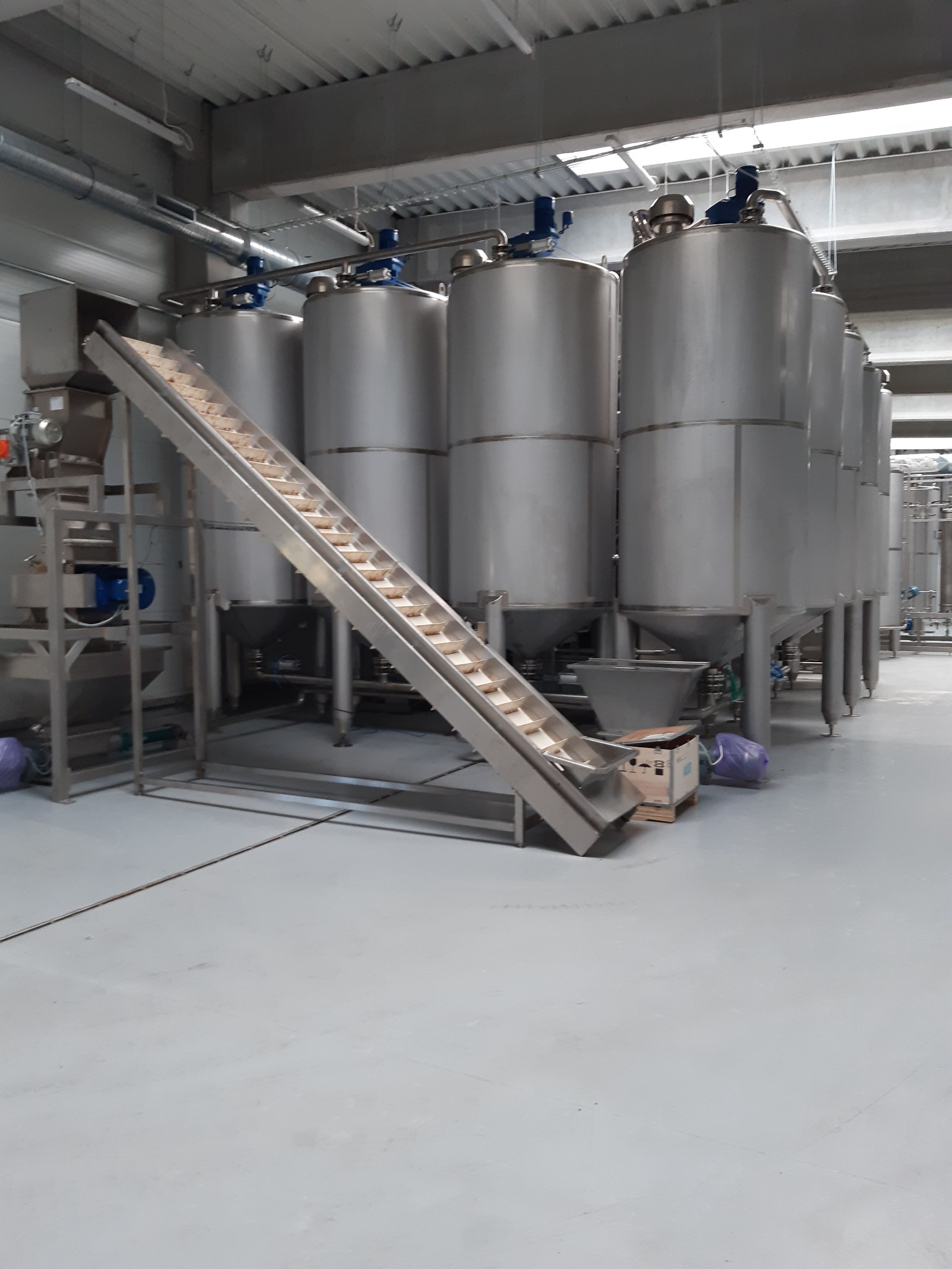 Metalowe silosy przemysłowe w hali produkcyjnej, z podajnikiem taśmowym transportującym produkty w pojemnikach.