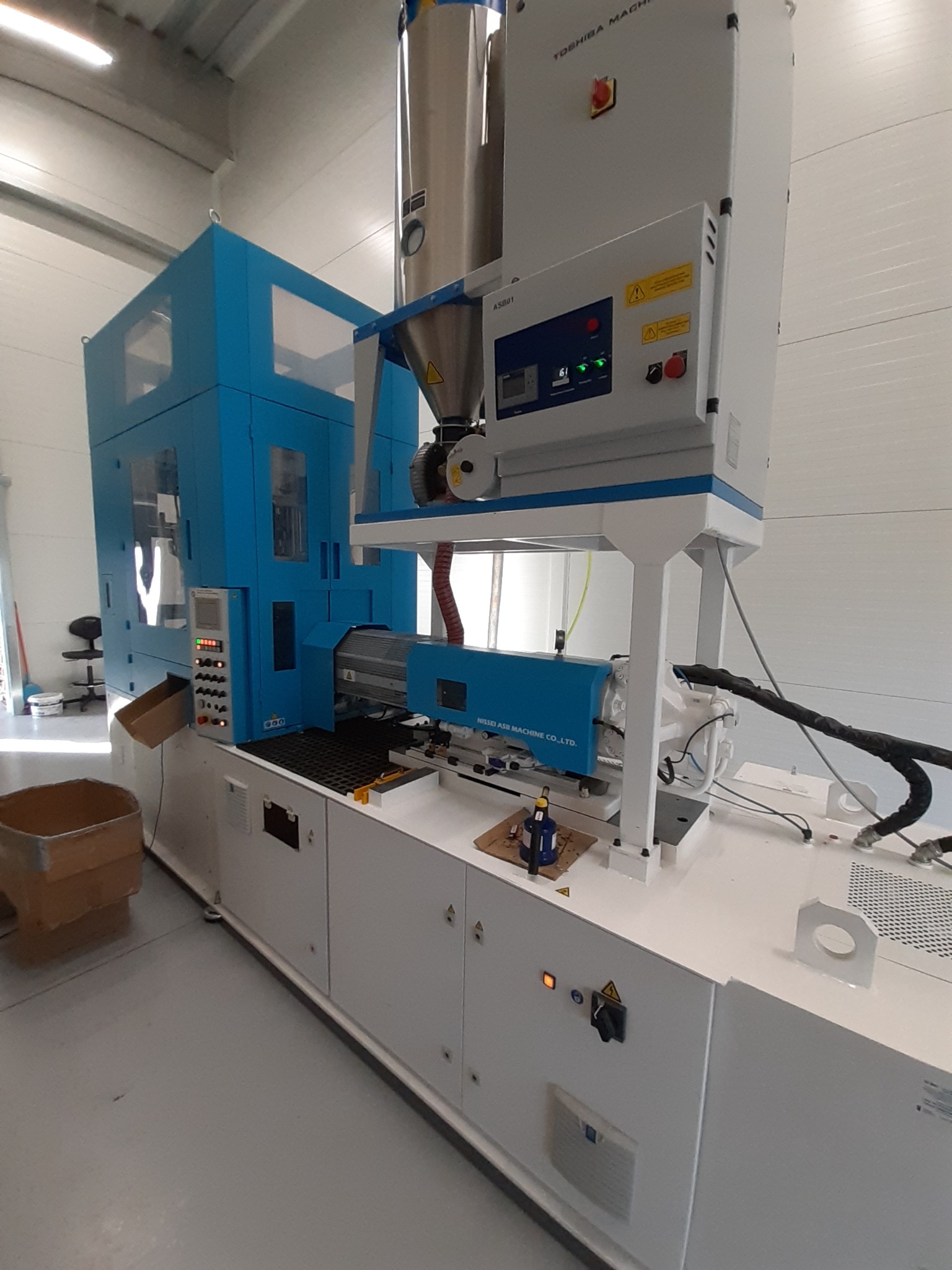 Maszyna do formowania wtryskowego tworzyw sztucznych marki Toshiba Machine i Nissei ASB Machine Co., Ltd. z widocznym panelem sterowania i zasobnikiem na granulat, umieszczona w hali produkcyjnej.