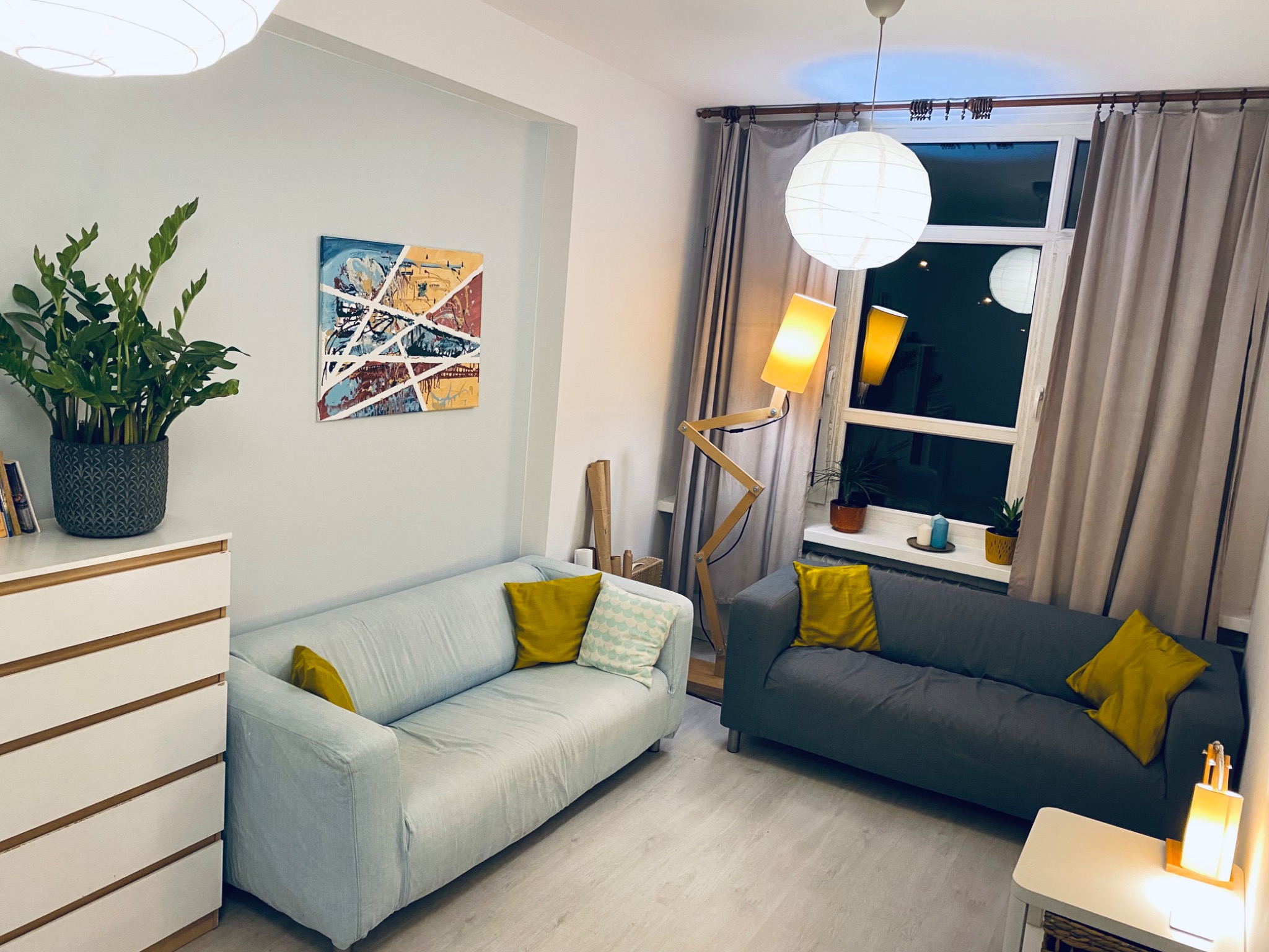 Jasny salon z dwoma sofami w odcieniach szarości i błękitu, zaakcentowany żółtymi poduszkami, z abstrakcyjnym obrazem na ścianie i lampą w kształcie robota z drewna.