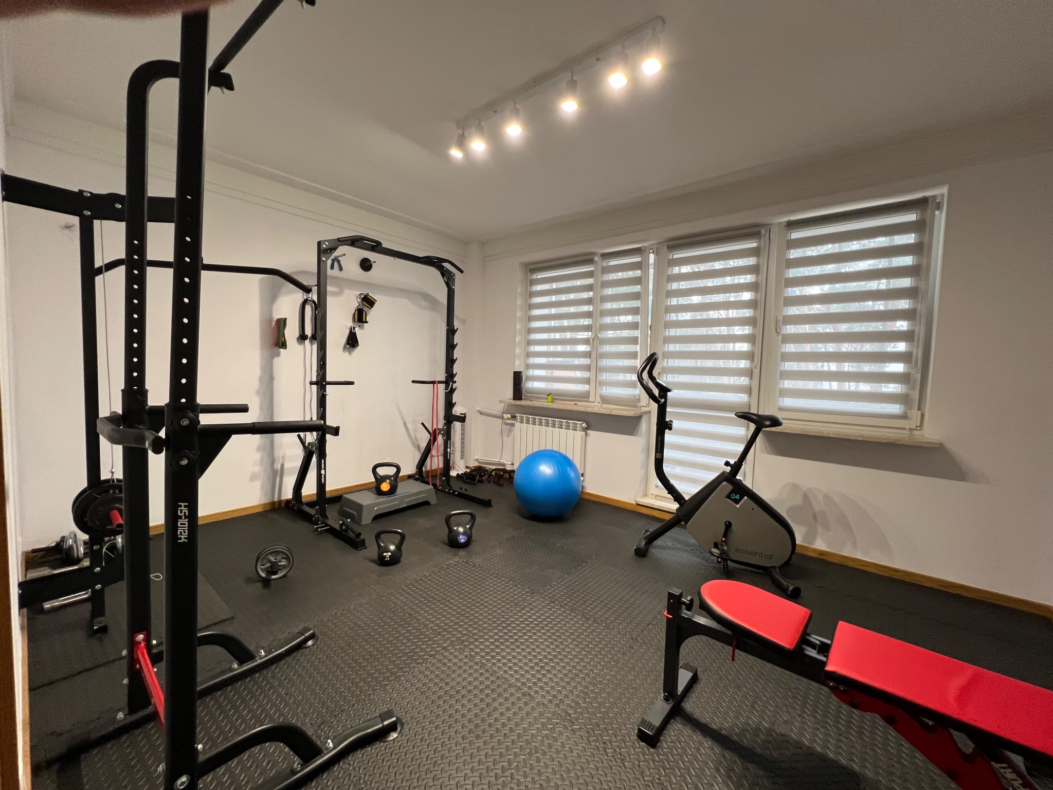 Domowa siłownia z czarną podłogą, wyposażona w atlas do ćwiczeń, sztangę, hantle kettlebell, piłkę gimnastyczną, rower stacjonarny i ławkę do ćwiczeń z czerwoną tapicerką, oświetlona listwą...