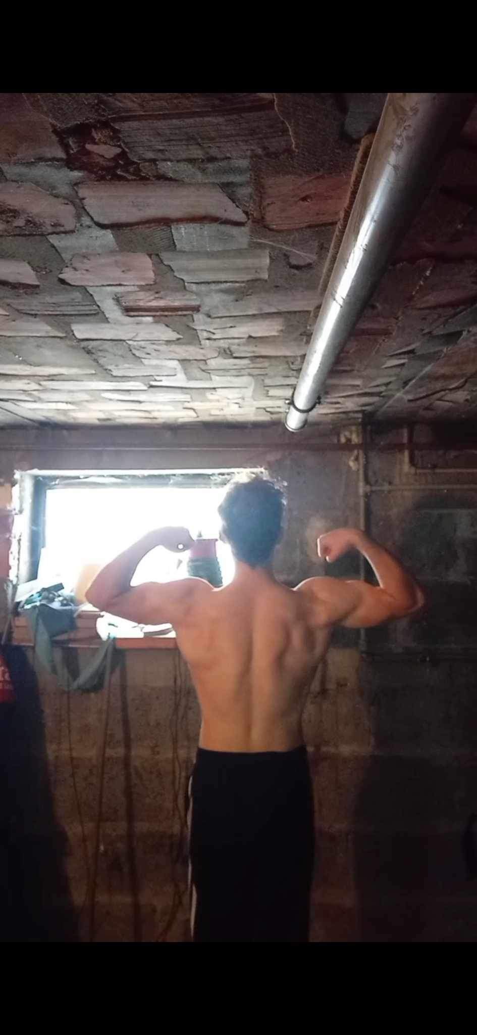 Mężczyzna prezentuje muskulaturę pleców i ramion, stojąc tyłem w piwnicy z oknem i odsłoniętą instalacją na suficie.