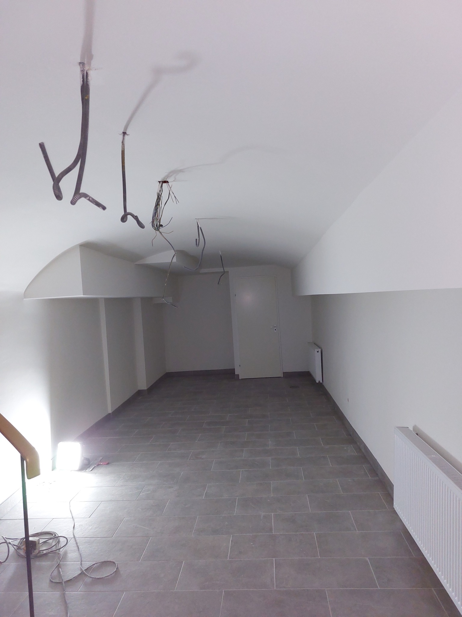 Wnętrze w trakcie wykończenia: szare płytki podłogowe, białe ściany i sufit, widoczne kable wystające z sufitu, grzejniki przy ścianach.