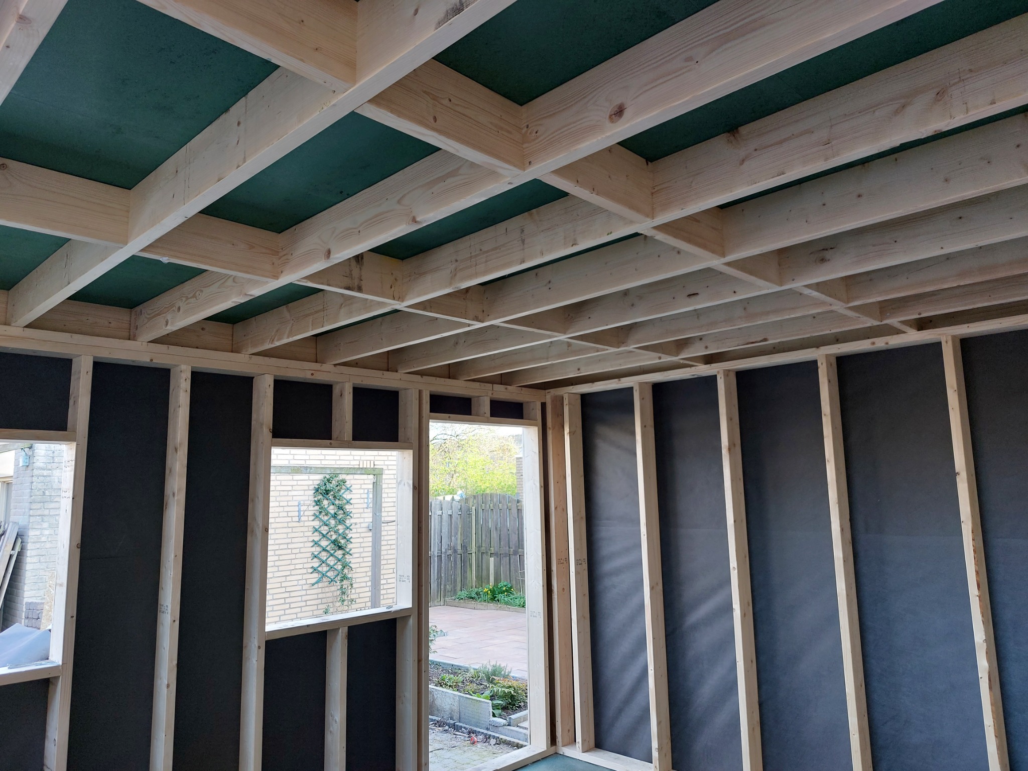 Wnętrze domu szkieletowego w trakcie budowy, widoczne drewniane ramy ścian, strop z zieloną płytą OSB oraz widok na ogród przez otwór okienny i drzwiowy.