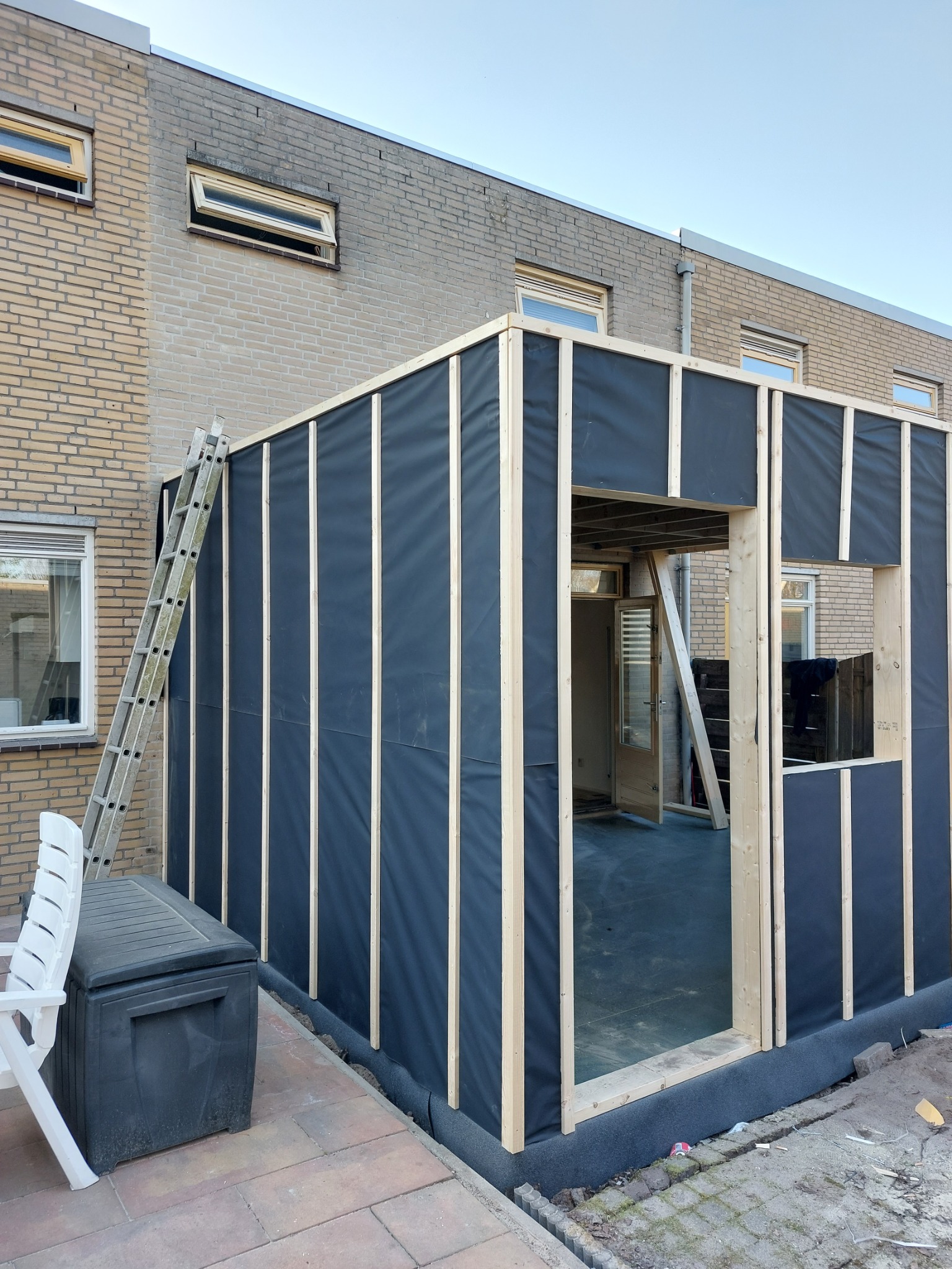 Konstrukcja szkieletowa dobudowy do domu jednorodzinnego z widoczną izolacją przeciwwilgociową, drewnianymi belkami, drabina oparta o ścianę budynku, skrzynia i krzesło na zewnątrz.