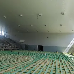 Przygotowanie podłoża pod parkiet w hali sportowej, widoczne drewniane legary ułożone na zielonej macie, pracownicy w tle, naturalne światło wpadające przez okna.