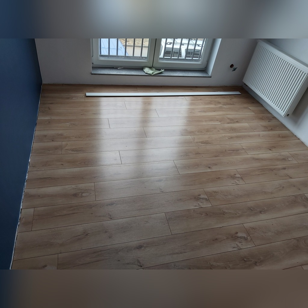 Jasne, drewnopodobne panele podłogowe ułożone w pokoju z niebieską ścianą i oknem, listwa przypodłogowa leży przy oknie.