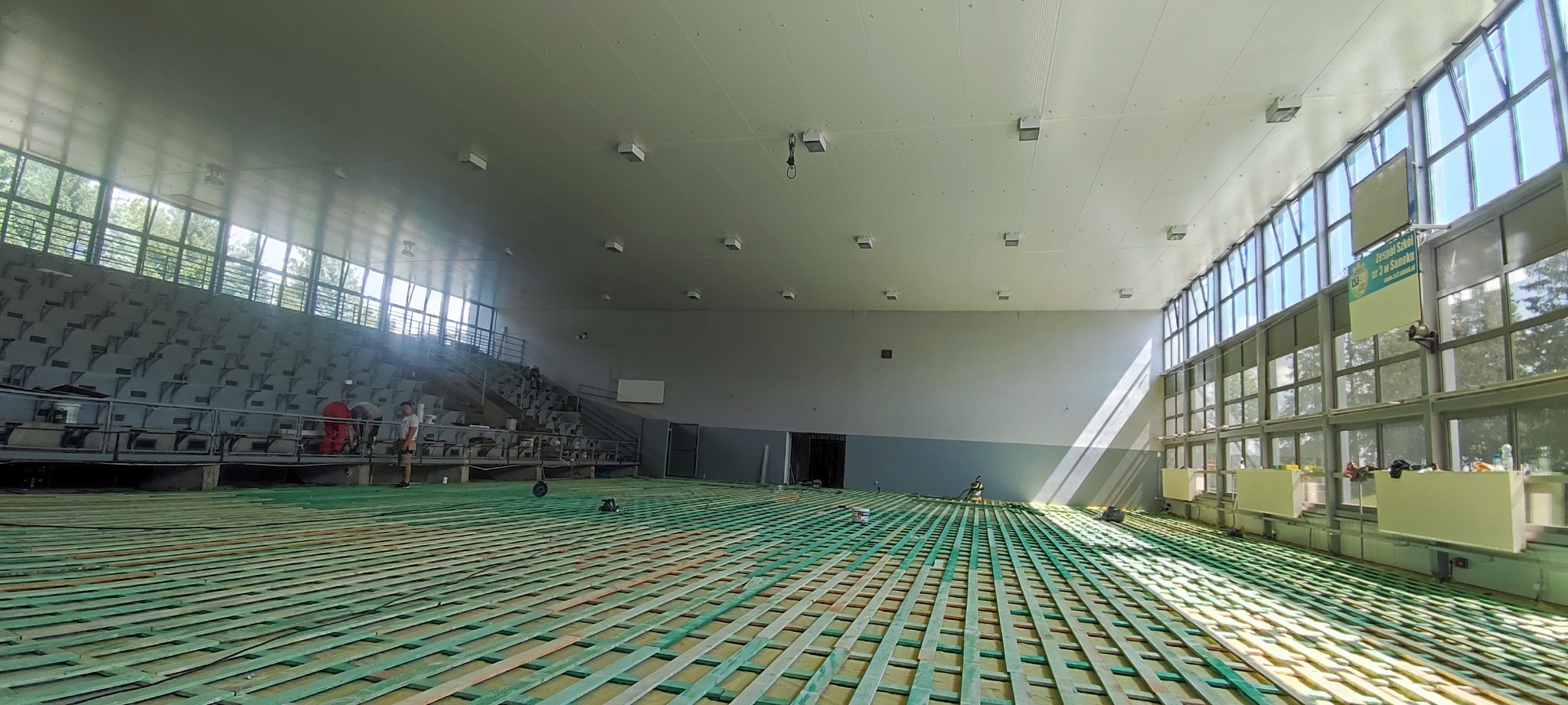 Przygotowanie podłoża pod parkiet w hali sportowej, widoczne drewniane legary ułożone na zielonej macie, pracownicy w tle, naturalne światło wpadające przez okna.