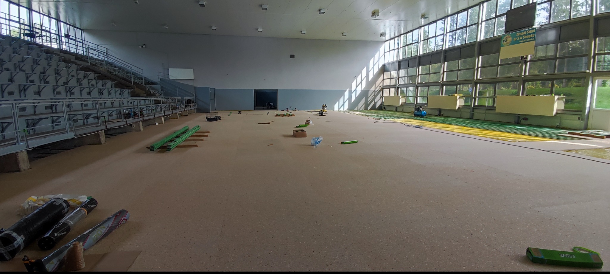 Sala gimnastyczna w trakcie remontu posadzki, widoczne rolki z materiałem podłogowym, narzędzia i częściowo ułożona podłoga w kolorach żółtym i zielonym, w tle trybuny i duże okna.