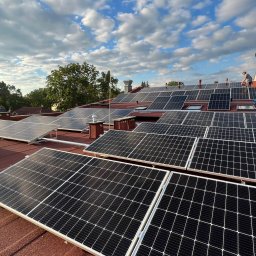 Wojciech Przepiórkowski PW CONTAKTON - Instalacja paneli słonecznych na czerwonym dachu domu jednorodzinnego, widoczny pracownik na drabinie, błękitne niebo z chmurami w tle.