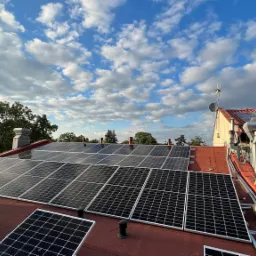 Instalacja paneli słonecznych na czerwonym dachu domu jednorodzinnego, widoczna drabina, butla gazowa i narzędzia montażowe, na tle błękitne niebo z chmurami.