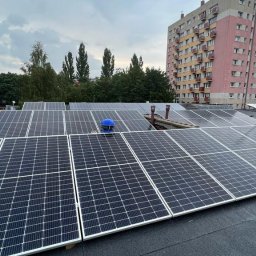 Wojciech Przepiórkowski PW CONTAKTON - Instalacja fotowoltaiczna na dachu bloku mieszkalnego, widoczny fragment niebieskiego kasku pracownika podczas montażu.