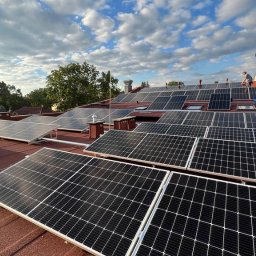 Wojciech Przepiórkowski PW CONTAKTON - Montaż paneli fotowoltaicznych na dachu domu z czerwoną dachówką, widoczne osoby pracujące na dachu, błękitne niebo z chmurami w tle.