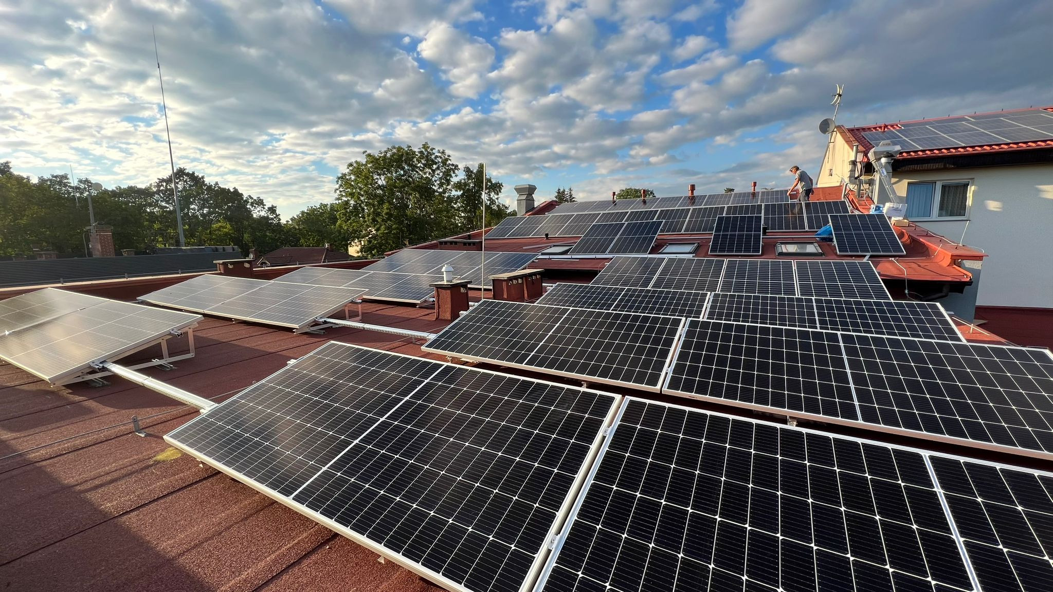 Instalacja paneli słonecznych na czerwonym dachu domu jednorodzinnego, widoczny pracownik na drabinie, błękitne niebo z chmurami w tle.