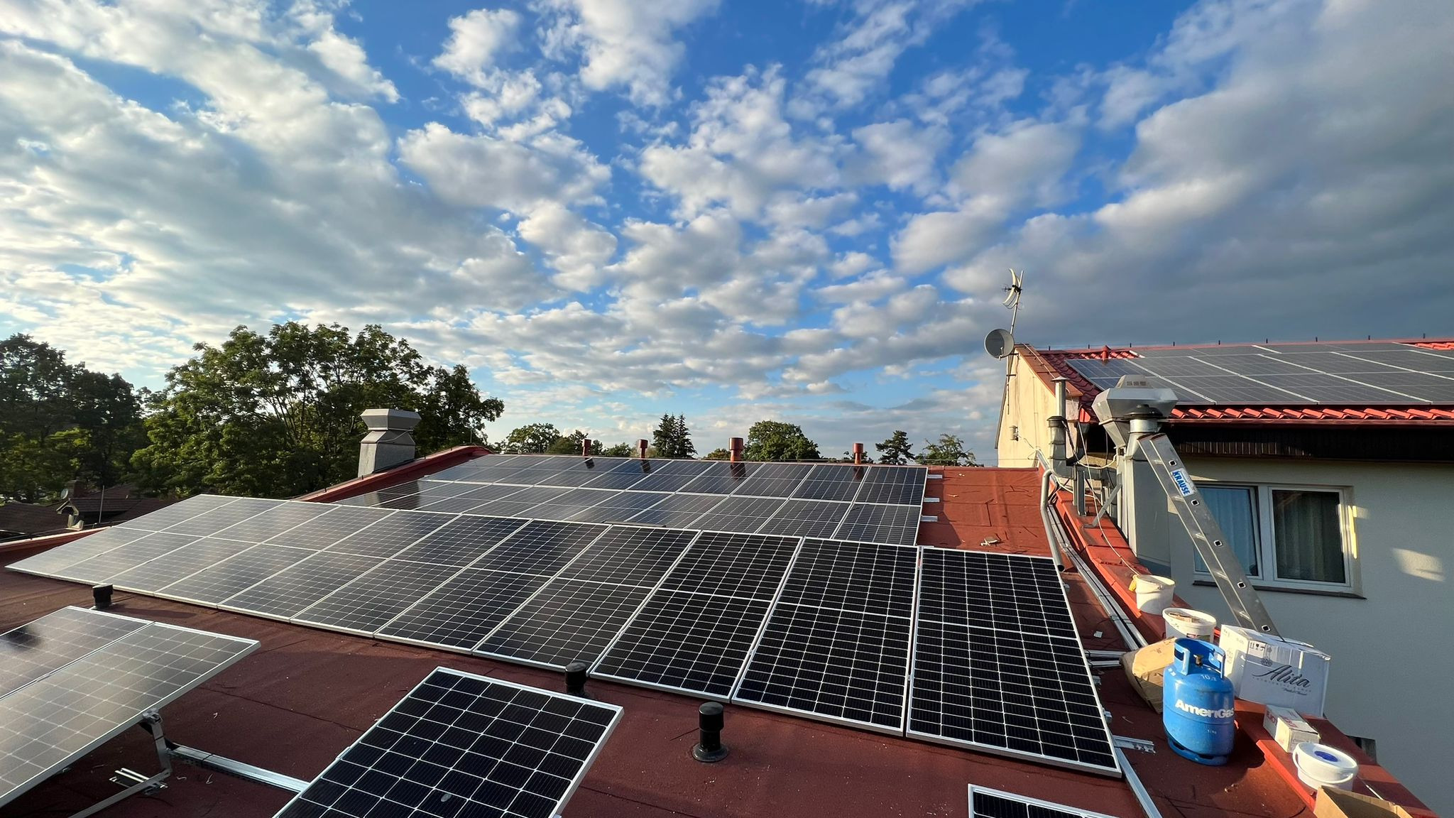 Instalacja paneli słonecznych na czerwonym dachu domu jednorodzinnego, widoczna drabina, butla gazowa i narzędzia montażowe, na tle błękitne niebo z chmurami.