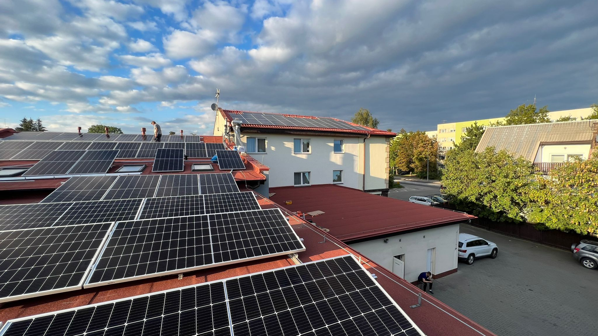 Instalacja fotowoltaiczna na dachu budynku z czerwonej dachówki, widok z góry na panele słoneczne, na dachu pracownik w jasnej koszuli, w tle budynki mieszkalne i błękitne niebo z chmurami.