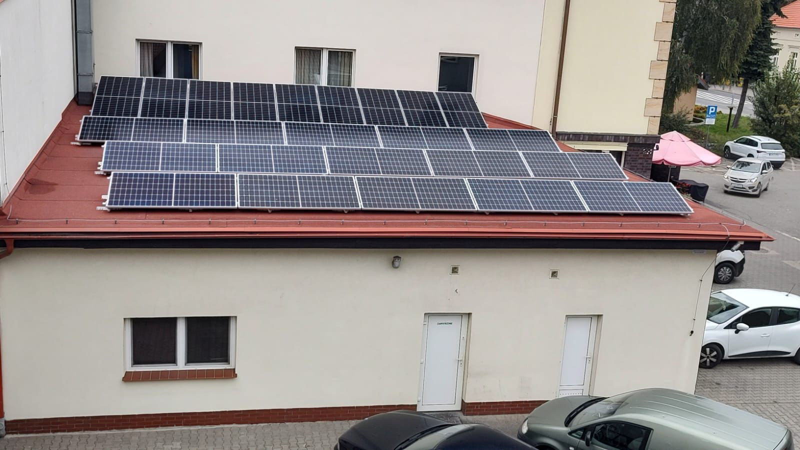 Panele słoneczne zamontowane na czerwonym dachu budynku, widoczne okna i drzwi na elewacji, a także zaparkowane samochody w otoczeniu.