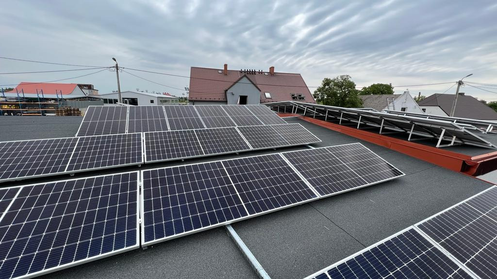 Instalacja fotowoltaiczna na dachu budynku z widocznymi panelami słonecznymi, elementami montażowymi i budynkami w tle pod pochmurnym niebem.