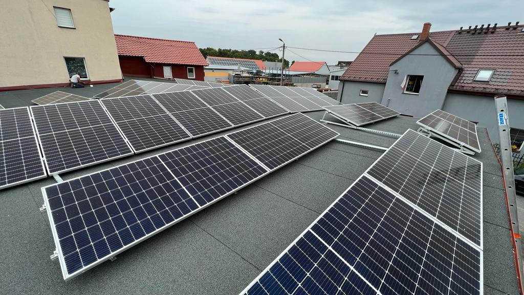 Montaż paneli słonecznych na płaskim dachu pokrytym papą, widoczna drabina Krause, w tle budynki mieszkalne z czerwonymi dachami.