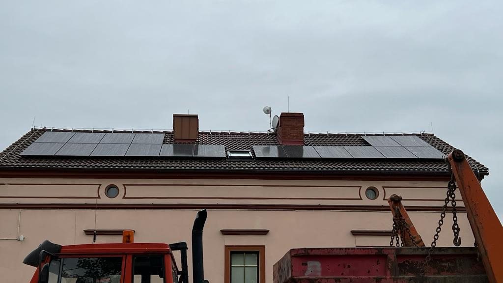 Częściowy widok instalacji paneli słonecznych na dachu budynku z ceglaną dachówką, z fragmentem pomarańczowego dźwigu budowlanego w dolnej części kadru, na tle pochmurnego nieba.