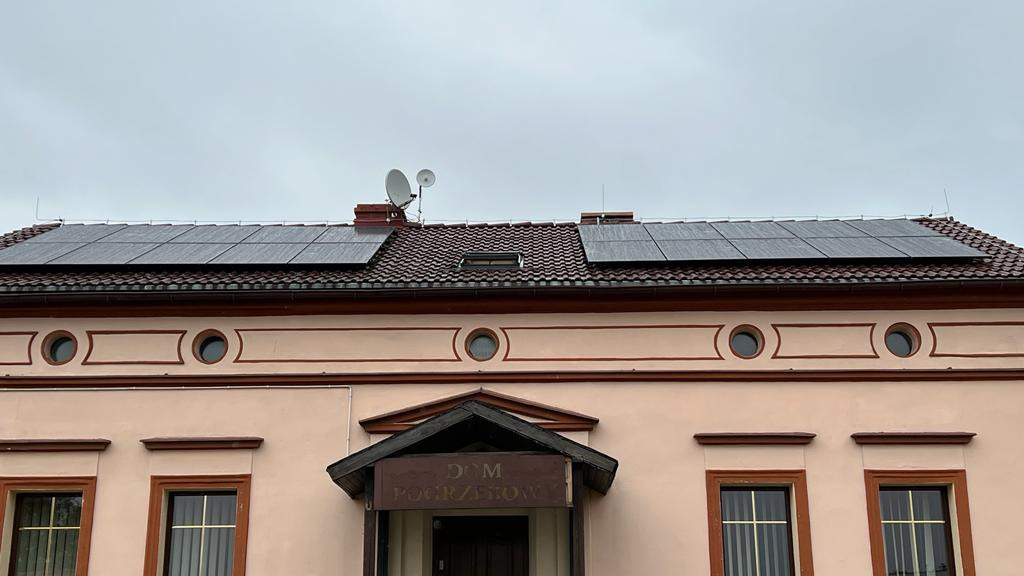 Panele słoneczne na dachu budynku z brązową dachówką, widoczne anteny satelitarne i odgromniki, budynek z jasną elewacją i oknami w brązowych ramach.