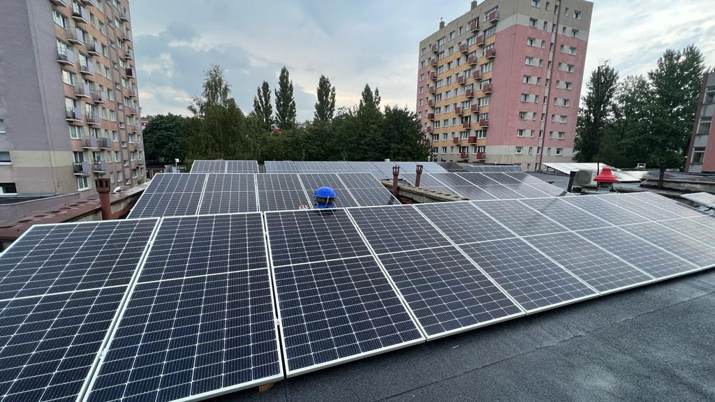 Instalacja fotowoltaiczna na dachu bloku mieszkalnego, widoczny fragment niebieskiego kasku pracownika podczas montażu.