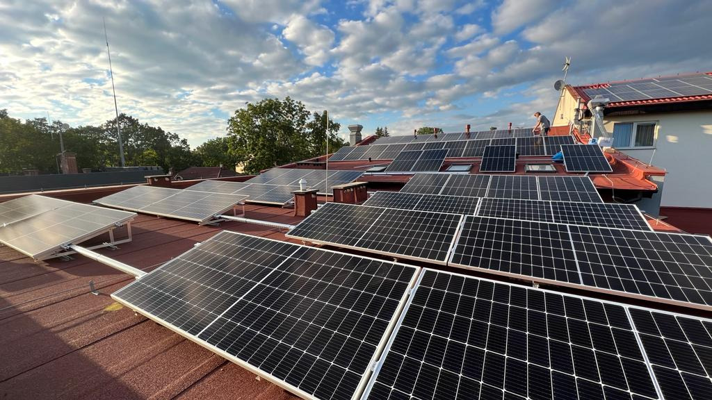 Montaż paneli fotowoltaicznych na dachu domu z czerwoną dachówką, widoczne osoby pracujące na dachu, błękitne niebo z chmurami w tle.