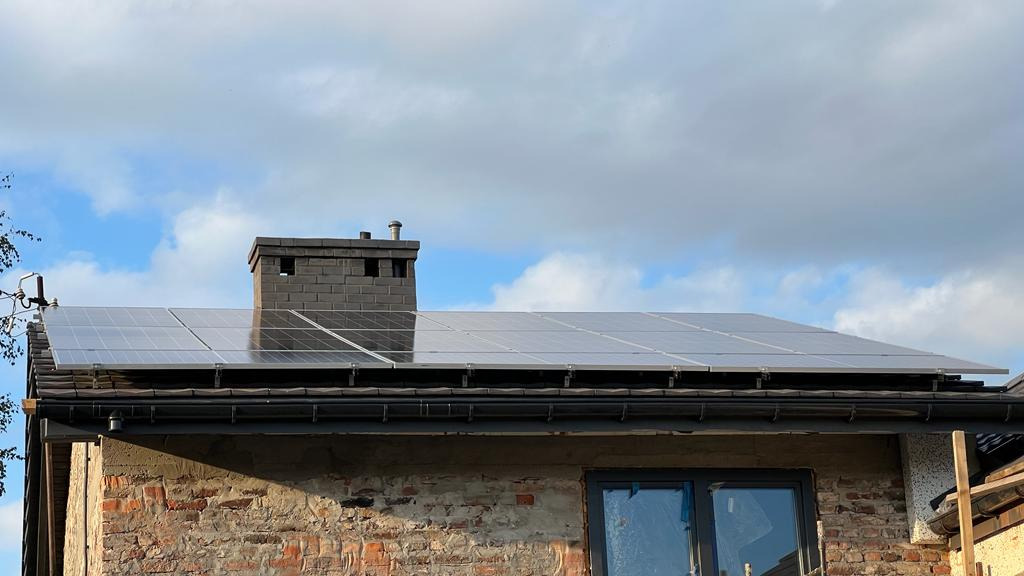 Panele słoneczne na dachu ceglanego domu z kominem, widoczne odbicia nieba na powierzchni paneli.